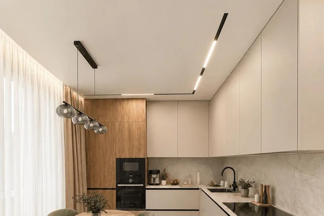 Светлая угловая кухня с обеденным столом, встроенным магнитным треком на матовом потолке и шторами, уходящими в нишу с подсветкой